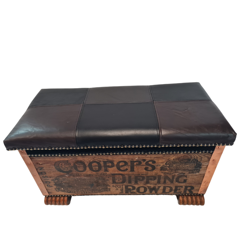 A vintage-style wooden storage chest or bench with a dark brown leather saddle cushion on top and a printed wood front panel reading 'Cooper's Dipping Powder.'.