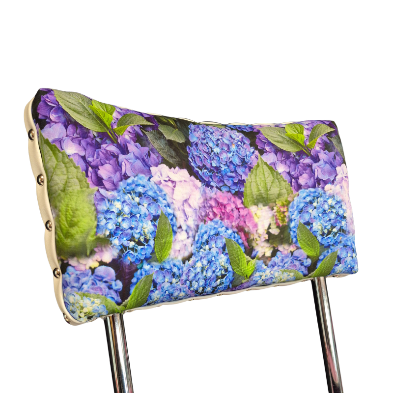 A close-up of a chair back cushion featuring a vibrant hydrangea floral print of blue, purple, and pink hydrangeas with green leaves. The cushion is secured with silver metal grommets to a light wood frame, with the chrome legs of the chair visible against a plain white background.