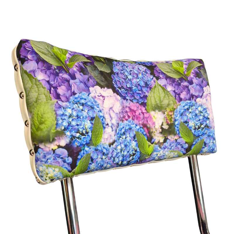 A close-up of a chair backrest cushion featuring a vibrant hydrangea floral print of blue, purple, and pink hydrangeas with green leaves, upholstered in a light beige fabric and supported by chrome metal legs.