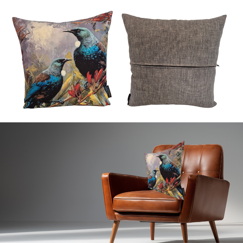 A decorative throw pillow shown in two views: a square printed pillow with two Tui's and NZ Flax on a muted background, and the reverse side in textured grey fabric. The bottom half shows the pillow styled on a brown leather armchair against a grey studio backdrop.